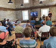 Staff member in a stone house in front of a projector screen giving a talk to a small room of people